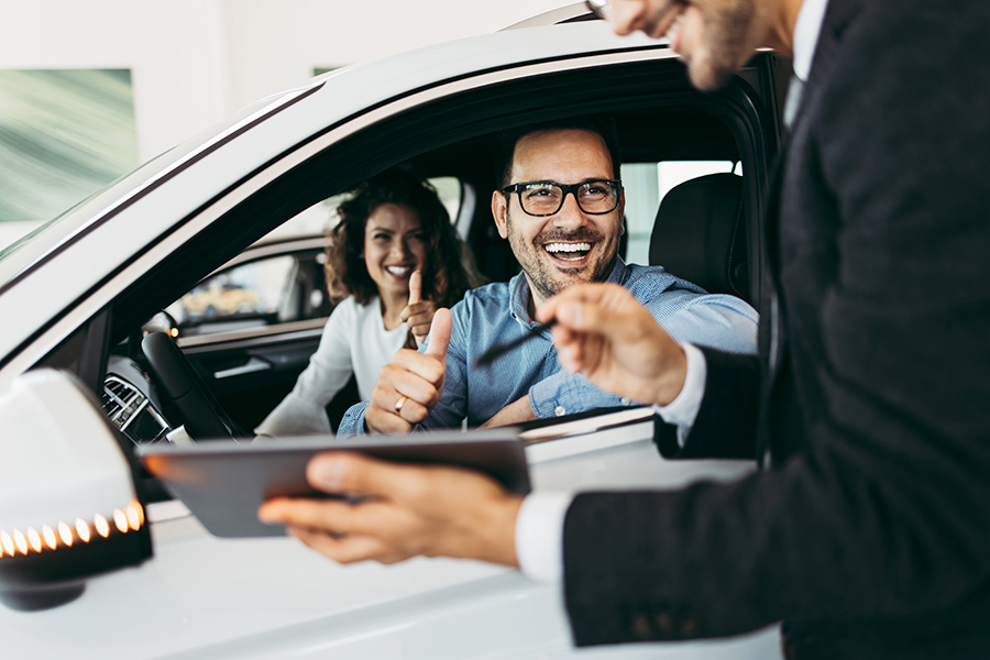 A smiling couple in their new GMC SUV give a thumbs-up to the smiling salesperson who's standing beside the vehicle