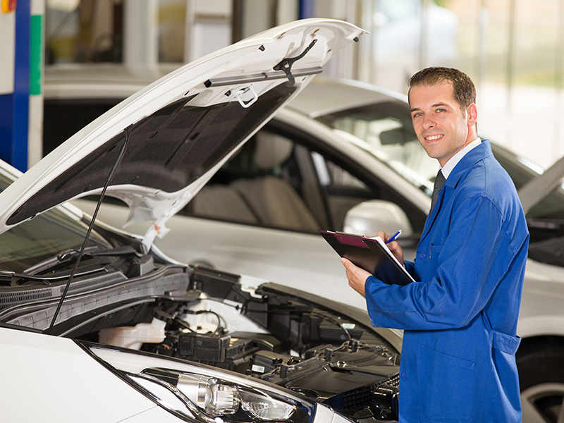 A smiling Certified Maintenance Professional checks off his task standing in front of the open car engine hood.