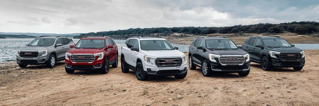 5 MODELS OF GMC SUVs lined up for show on the sand beside a lake with low hills and a blue-grey sky behind